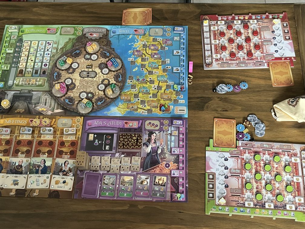 Das Bild zeigt das Brettspiel Ada's Dream. Es ist auf einem Tisch aufgebaut, mit mehreren Spielbrettern und Komponenten, die sorgfältig platziert sind. Zu sehen sind verschiedene Bereiche des Spiels: Links befindet sich das "Institutions"-Spielbrett, darüber das "Workshop"-Brett, das eine Art zentralen Bereich darstellt, in dem Würfel und Marker verwendet werden. Im mittleren Bereich des Spiels wird das "Travel"-Brett gezeigt, auf dem Karten und Marker abgebildet sind. Rechts sieht man das "Ada's Study"-Brett, das den Forschungsbereich zeigt, und darunter das "Meetings"-Brett, in dem Arbeitskarten und Ressourcen verwaltet werden. Auf dem Tisch liegen außerdem diverse Marker und Spielsteine in verschiedenen Farben, sowie einige verdeckte Karten, die noch nicht gezogen wurden.