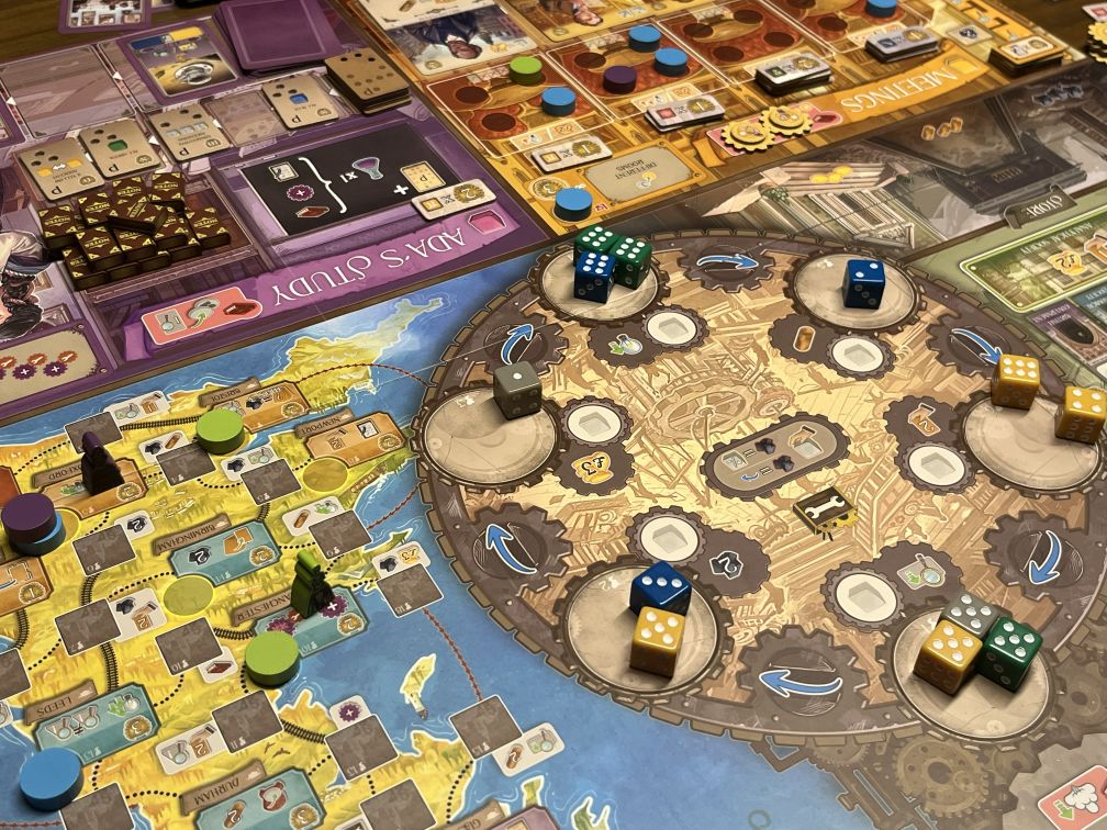 Das Bild zeigt einen Ausschnitt aus dem Brettspiel Ada's Dream. Im Mittelpunkt steht das zentrale "Workshop"-Spielfeld, auf dem verschiedene Würfel in verschiedenen Farben (grün, blau, gelb) platziert sind. Diese Würfel sind Teil des Aktionsmechanismus des Spiels und befinden sich auf Zahnrädern, die sich drehen. Links sieht man das "Ada's Study"-Spielfeld mit einigen Karten und Ressourcen, die darauf platziert sind. Auf dem unteren Teil des Spiels ist das "Travel"-Brett zu sehen, auf dem Spielfiguren (in Form von kleinen Pöppeln) und Marker entlang eines Routenplans bewegen. Die Spielkomponenten sind in lebendigen Farben und zeigen den aktuellen Stand des Spiels.