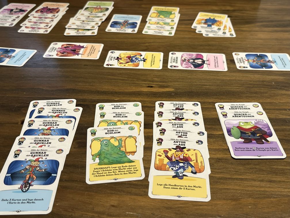 Auf einem Holztisch liegt eine ausgebreitete Spielsituation aus Die glorreichen Gilden von Buttonville. Mehrere Reihen von Karten mit bunten Illustrationen und Charakteren sind zu Sets geordnet, etwa „Gunnar der Gaukler“, „Bob der Schleim“, „Astor das Ass“ und „Quacks der Abenteurer“. Im Hintergrund sind weitere Kartenstapel verteilt, die auf ein laufendes Spiel hindeuten. Die lebhaften Farben und Figuren zeigen eine humorvolle Fantasy-Atmosphäre.