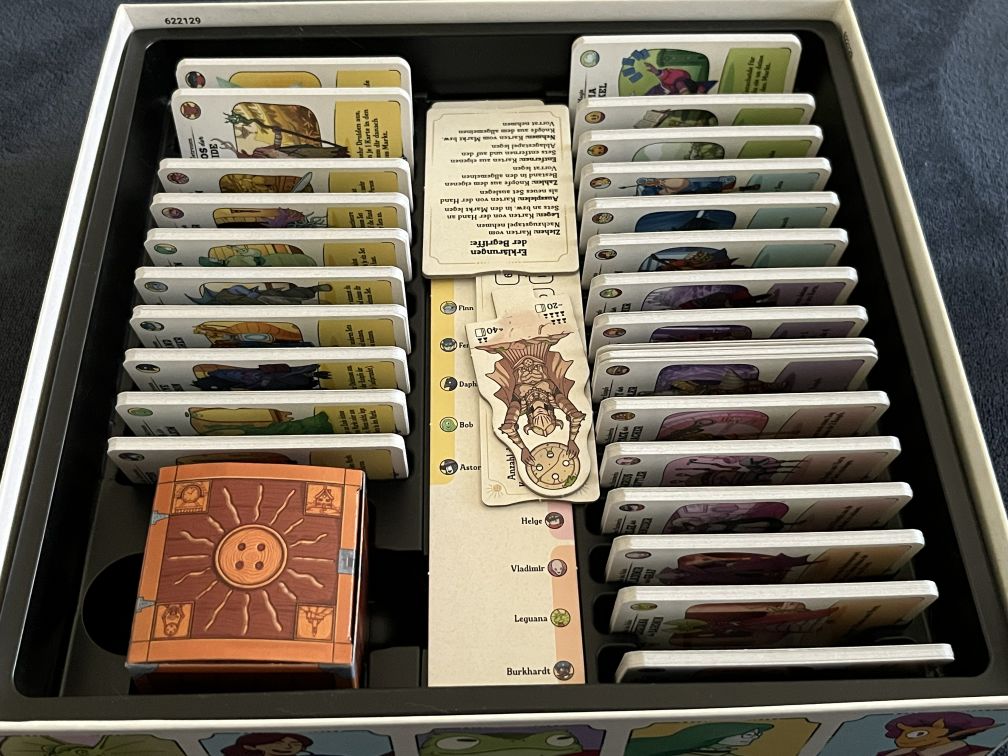 Geöffneter Spielekarton des Brettspiels Die glorreichen Gilden von Buttonville. Im Einsatzfach liegen ordentlich sortierte Kartenstapel mit verschiedenen Charakteren und Illustrationen in bunten Farben. Links unten befindet sich eine braune, würfelförmige Schachtel mit einem Knopf-Symbol in der Mitte, daneben mehrere Reihen von Karten und Anleitungen. Die Karten zeigen unterschiedliche Figuren aus den Gilden, wie etwa Abenteurer, Geister und Handwerker.