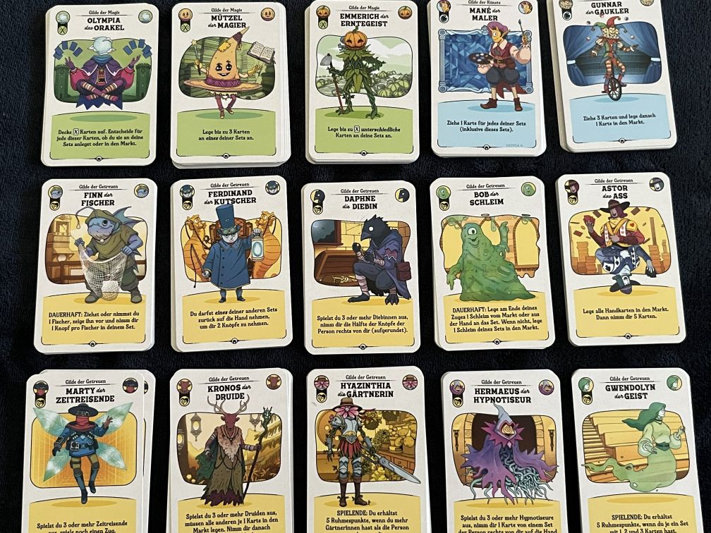 Auf einem dunklen Stoff liegen fünfzehn Spielkarten aus Die glorreichen Gilden von Buttonville in drei Reihen angeordnet. Jede Karte zeigt eine bunte Illustration eines Charakters mit Namen, Gilde und Spieltext. Oben befinden sich Figuren wie „Olympia das Orakel“ und „Gunnar der Gaukler“, in der Mitte Charaktere wie „Finn der Fischer“ und „Bob der Schleim“, unten unter anderem „Marty der Zeitreisende“, „Kronos der Druide“ und „Gwendolyn der Geist“. Die Karten sind farblich und thematisch unterschiedlich gestaltet.