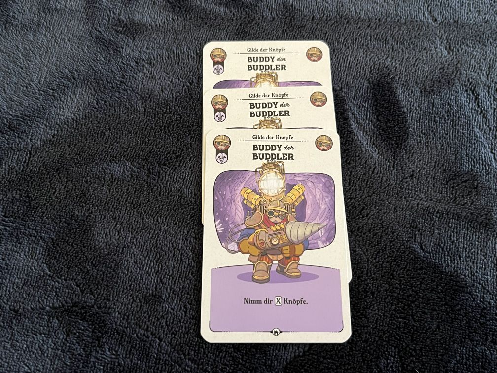 Drei Spielkarten aus dem Brettspiel Die glorreichen Gilden von Buttonville liegen gestapelt auf einem dunklen Stoffuntergrund. Die oberste Karte zeigt die Figur „Buddy der Buddler“ in einem gelben Bohranzug mit großem Bohrer und Helmlicht vor einem lilafarbenen Hintergrund. Unten auf der Karte steht der Text „Nimm dir X Knöpfe.“